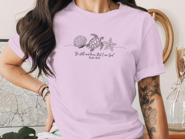 Purple t-shirt with minimalist line art of seashell, starfish, and turtle with "Be still and know that I am God" text.