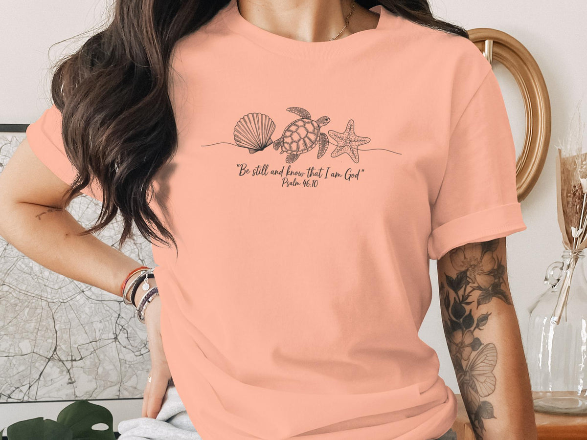 Peach t-shirt with a line drawing of a seashell, starfish, and sea turtle above Bible verse.