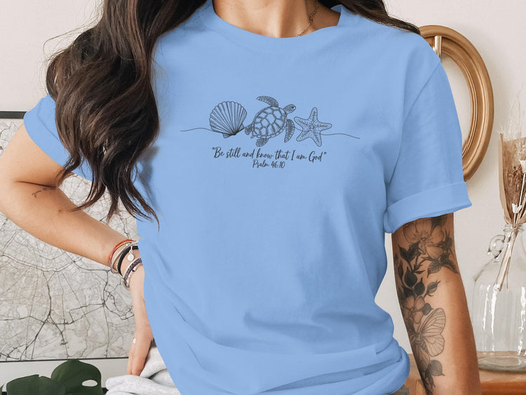 Light blue t-shirt with line art of seashell, starfish, and turtle, and text "Be still and know that I Am God Psalm 46:10".