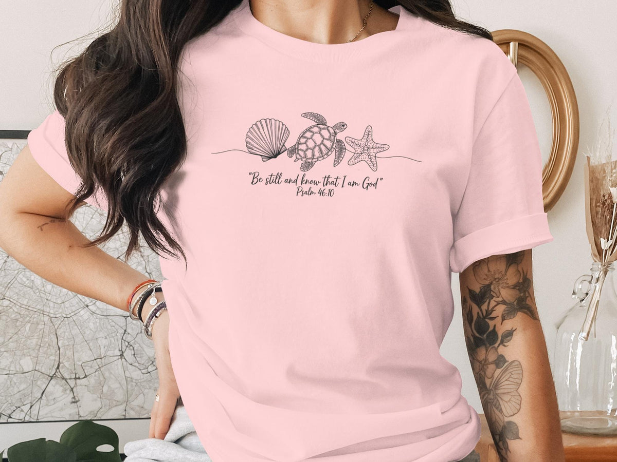 Pink t-shirt with line art of seashell, starfish, and turtle, and text "Be still and know that I am God"