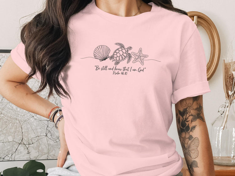 Pink t-shirt with line art of seashell, starfish, and turtle, and text "Be still and know that I am God"