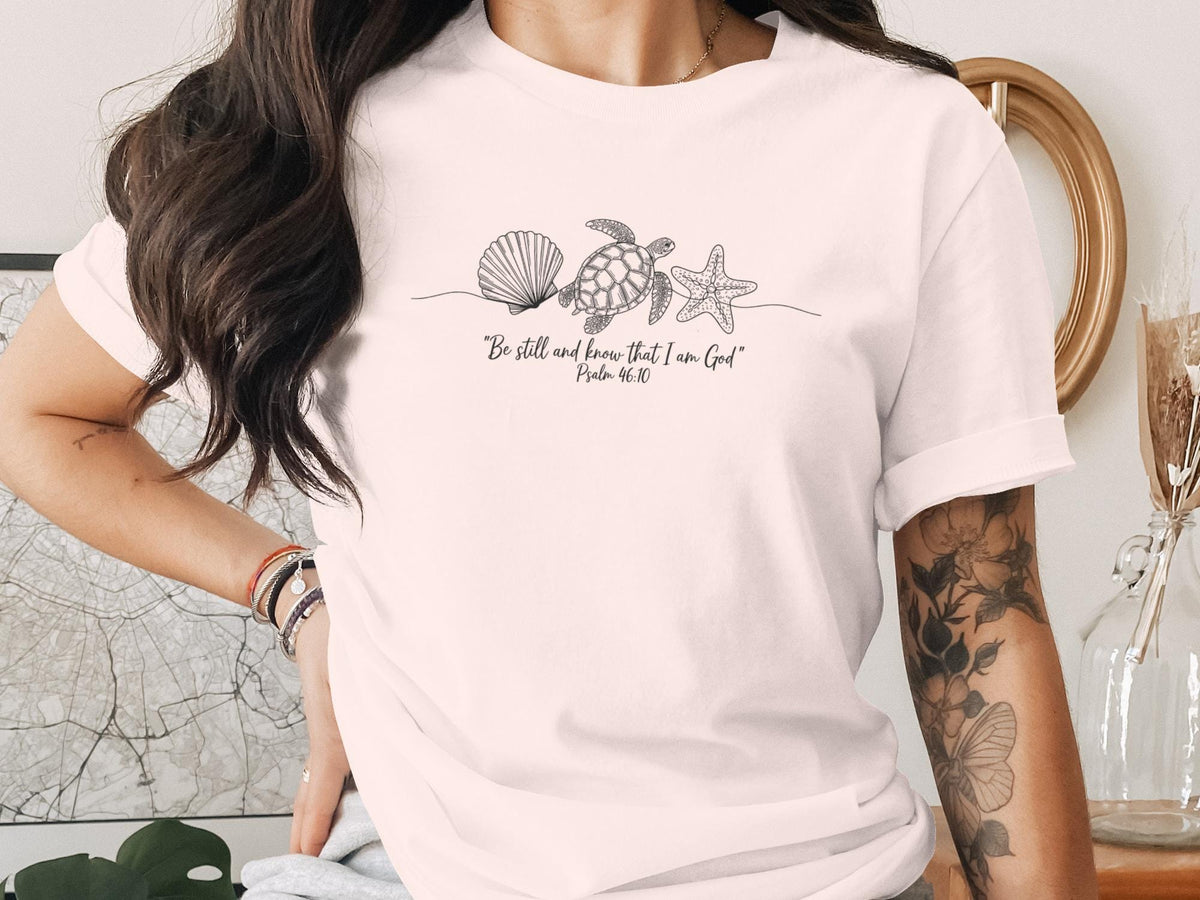 White t-shirt with line art of a seashell, starfish, and turtle, and text "Be still and know that I am God. Psalm 46:10".