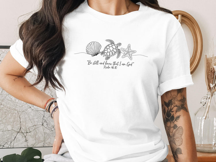 White t-shirt with black line art of a seashell, starfish, and turtle, and text "Be still and know that I am God".