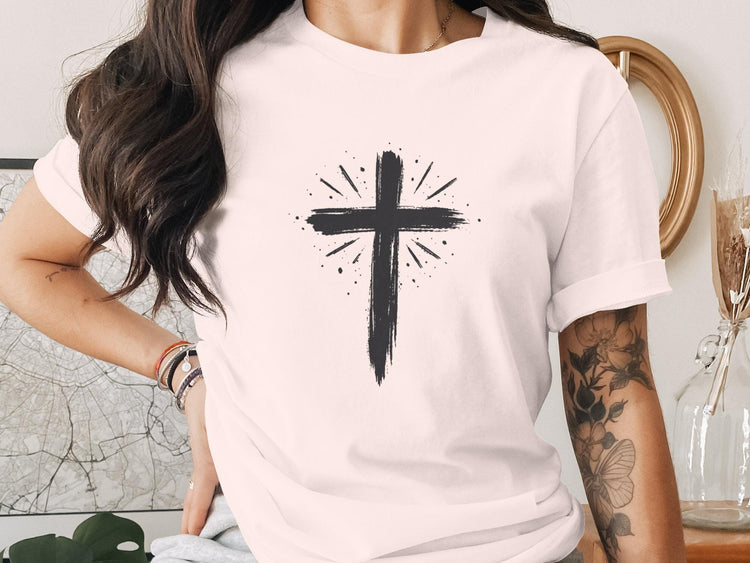 Black brushstroke cross with radiating lines on a light pink t-shirt.