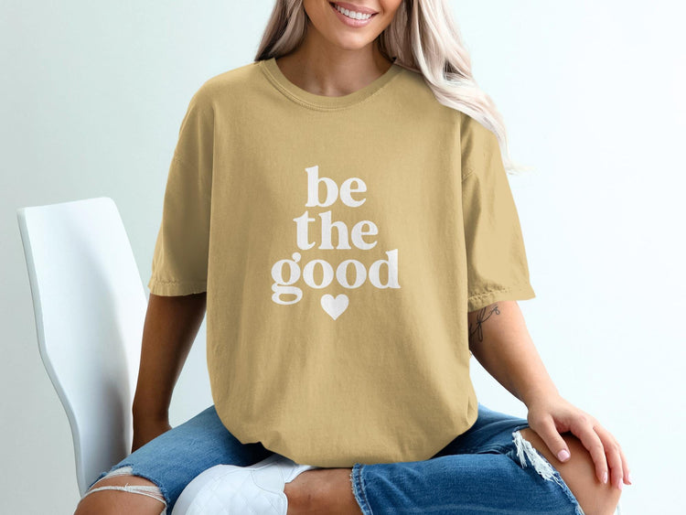 Tan t-shirt with white text "be the good" and a heart graphic.