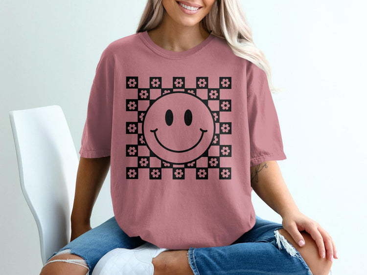 A person wearing a dusty pink t-shirt printed with a black smiley face and checkered flower pattern.