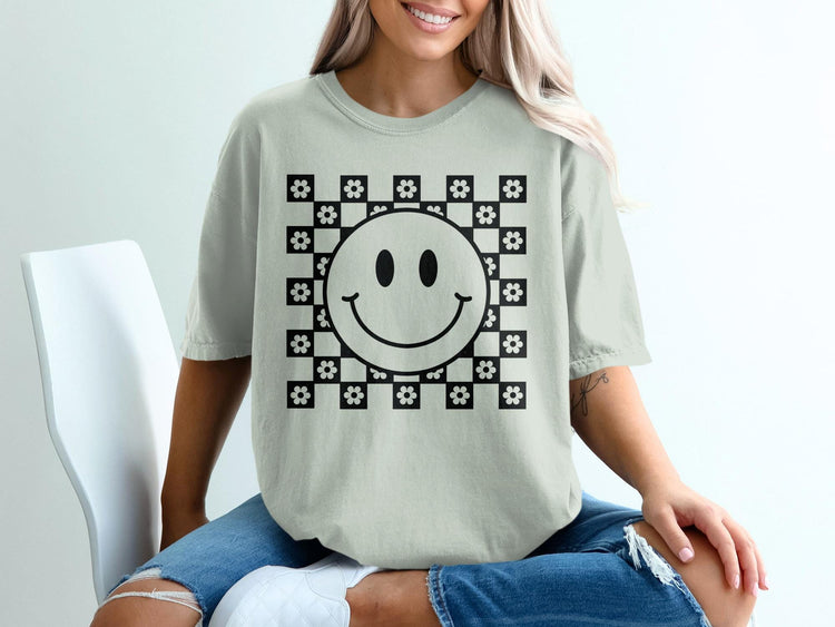 A woman wearing a light gray t-shirt featuring a black and white checkered and smiley face graphic.