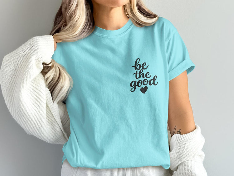 Light blue t-shirt with black script text "be the good" and a small heart.