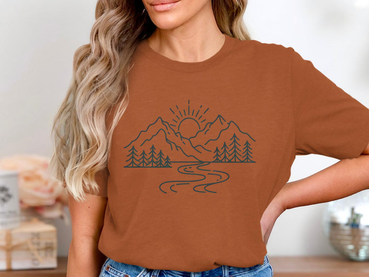 Brown t-shirt with a minimalist line art design of mountains, trees, and a river at sunrise.