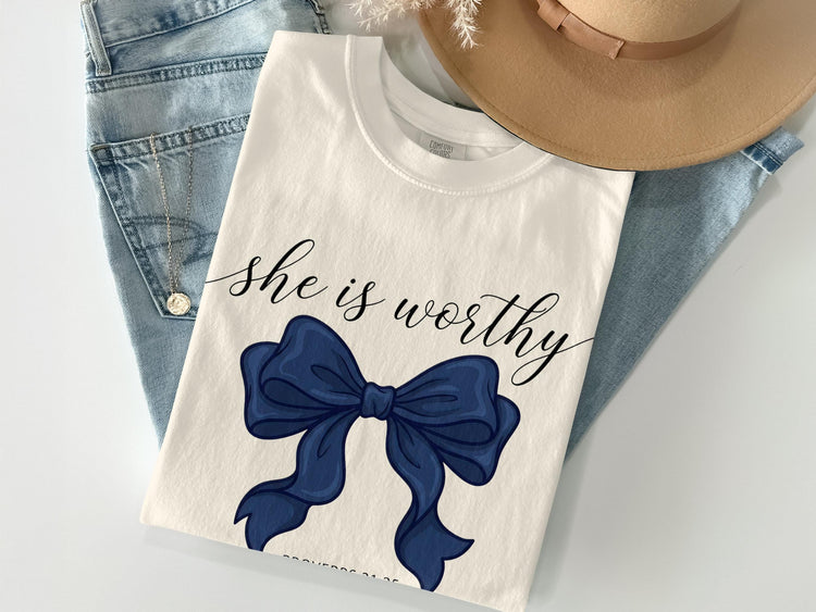 White t-shirt with "she is worthy" text and a blue bow, laid on jeans.