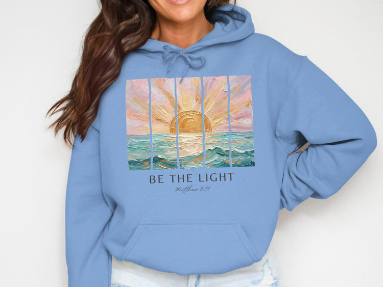 Light blue hoodie with sunset art and text "BE THE LIGHT Matthew 5:14".